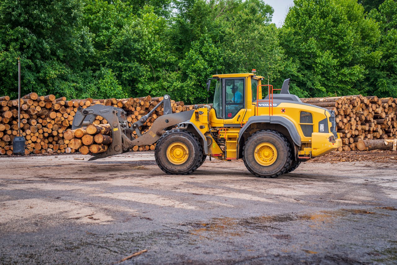 A yellow tractor lifting tree logs in an outdoor timber yard, surrounded by greenery.